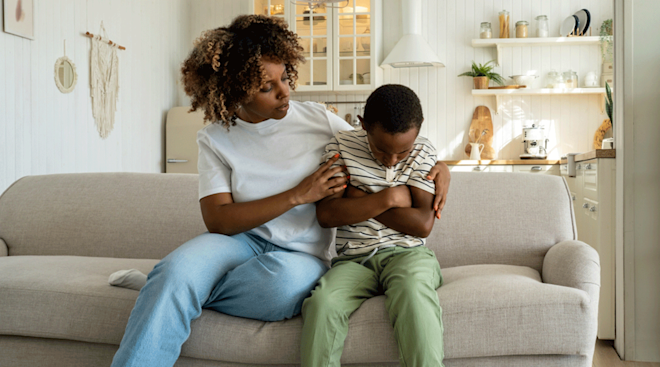 Young boy sitting on the couch with his arms folded in anger, with his caregiver providing comfort.
